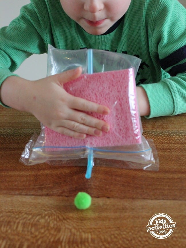 Demonstrate air pressure experiment for kids - baggie with sponges and straw propelling pom pom