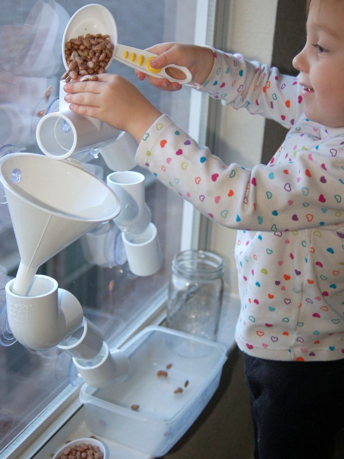 Tube Play- child pouring beans into the window stuck pvc tubes and funnels- kids activities blog