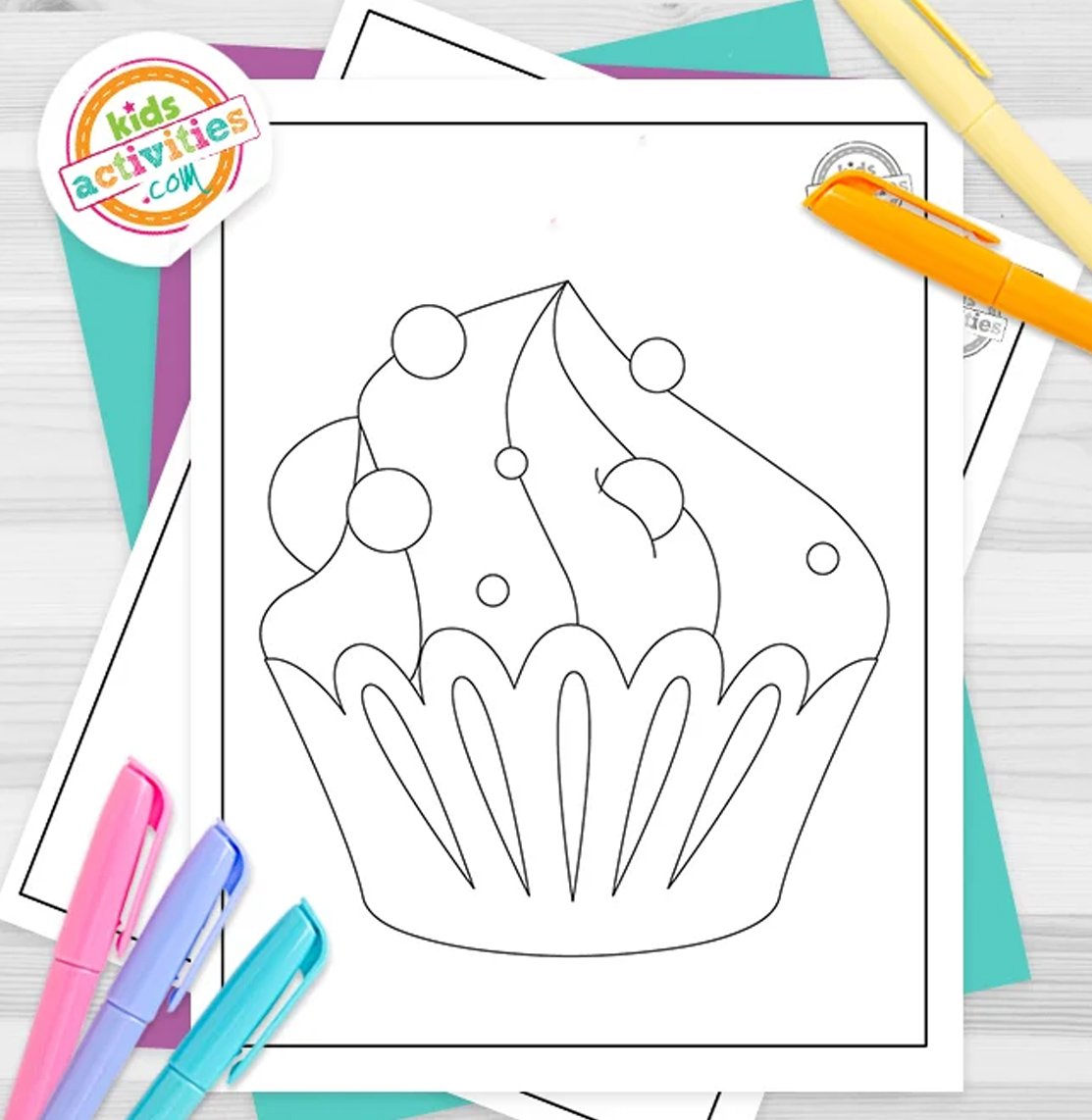 Stack of printed cupcake coloring page printables on top of green and purple pages, with markers next to them, on top of a gray background.