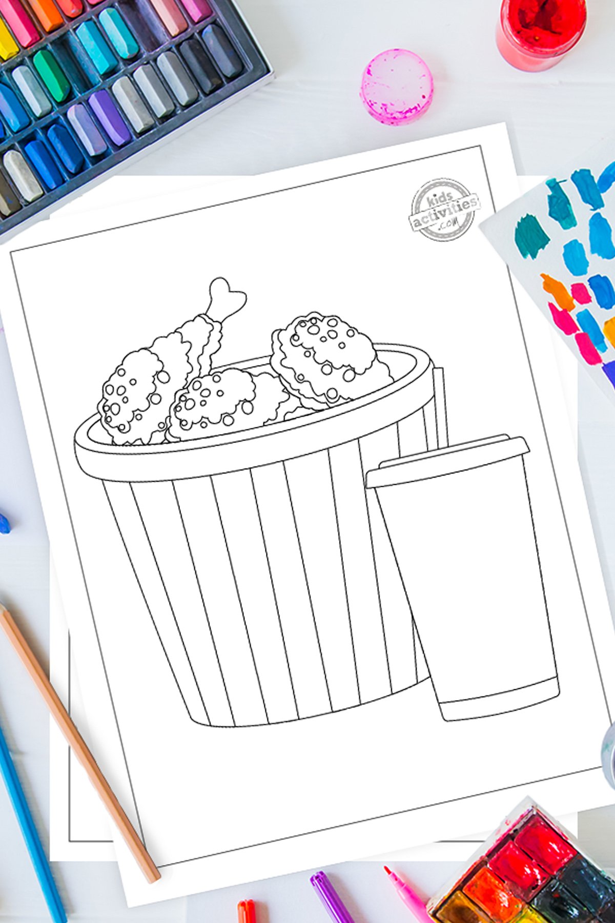 Black and white fried chicken coloring pages lying on top of blue-green and purple sheets with assorted crayons on a gray background.