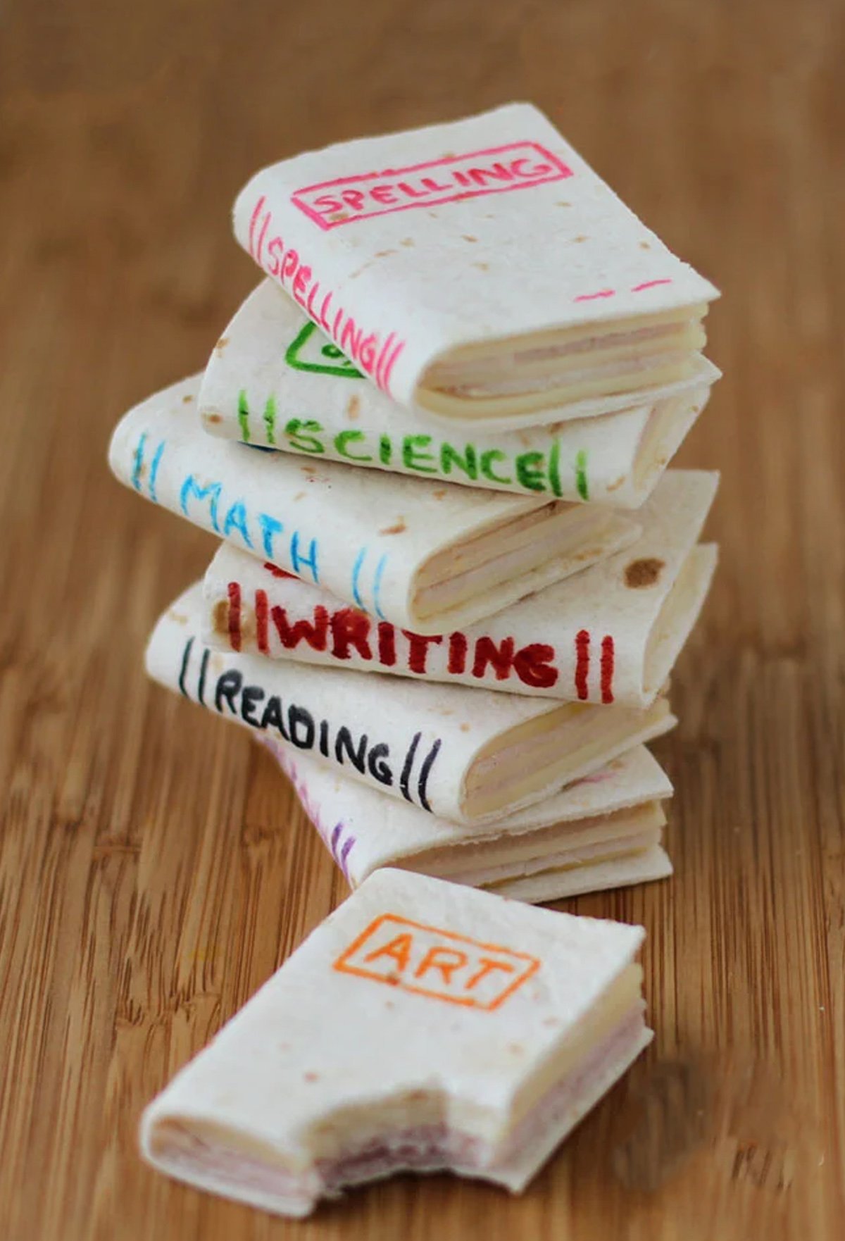 A stack of book-shaped mini sandwiches with different school subjects written on them, like Art, Reading, writing, Math, and Science.