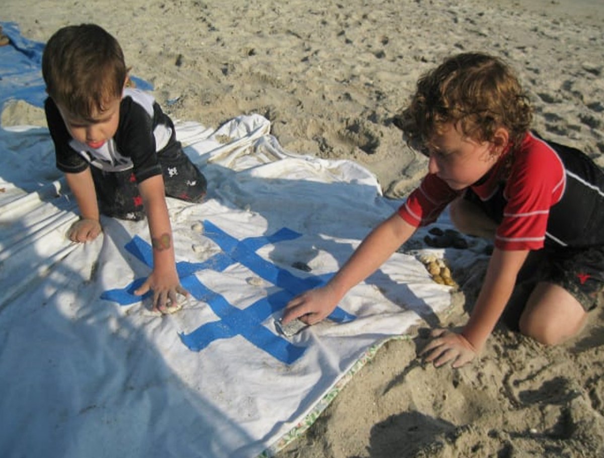 Two kids playing tic-tac-toe on a mat on a beach.