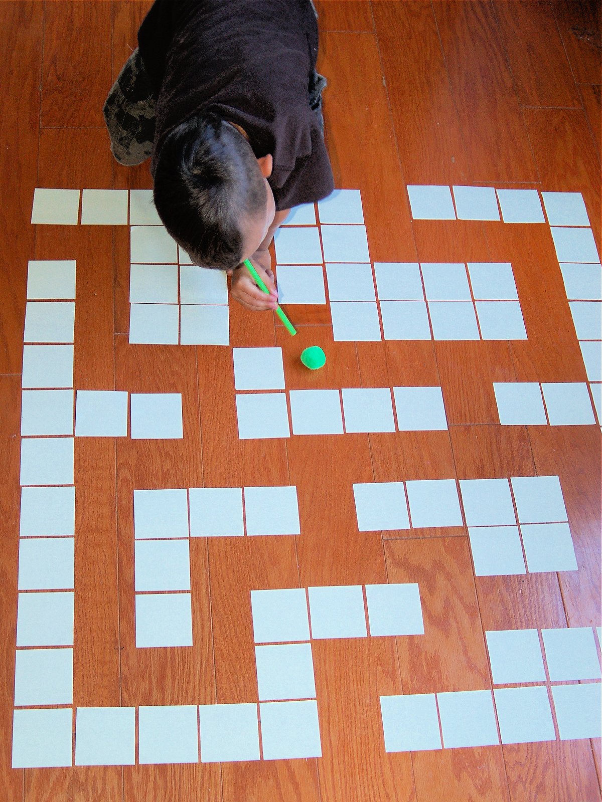 DIY Maze- completed- little boy using a green straw to blow a green pom pom along a paper maze on a wood floor- kids activities blog