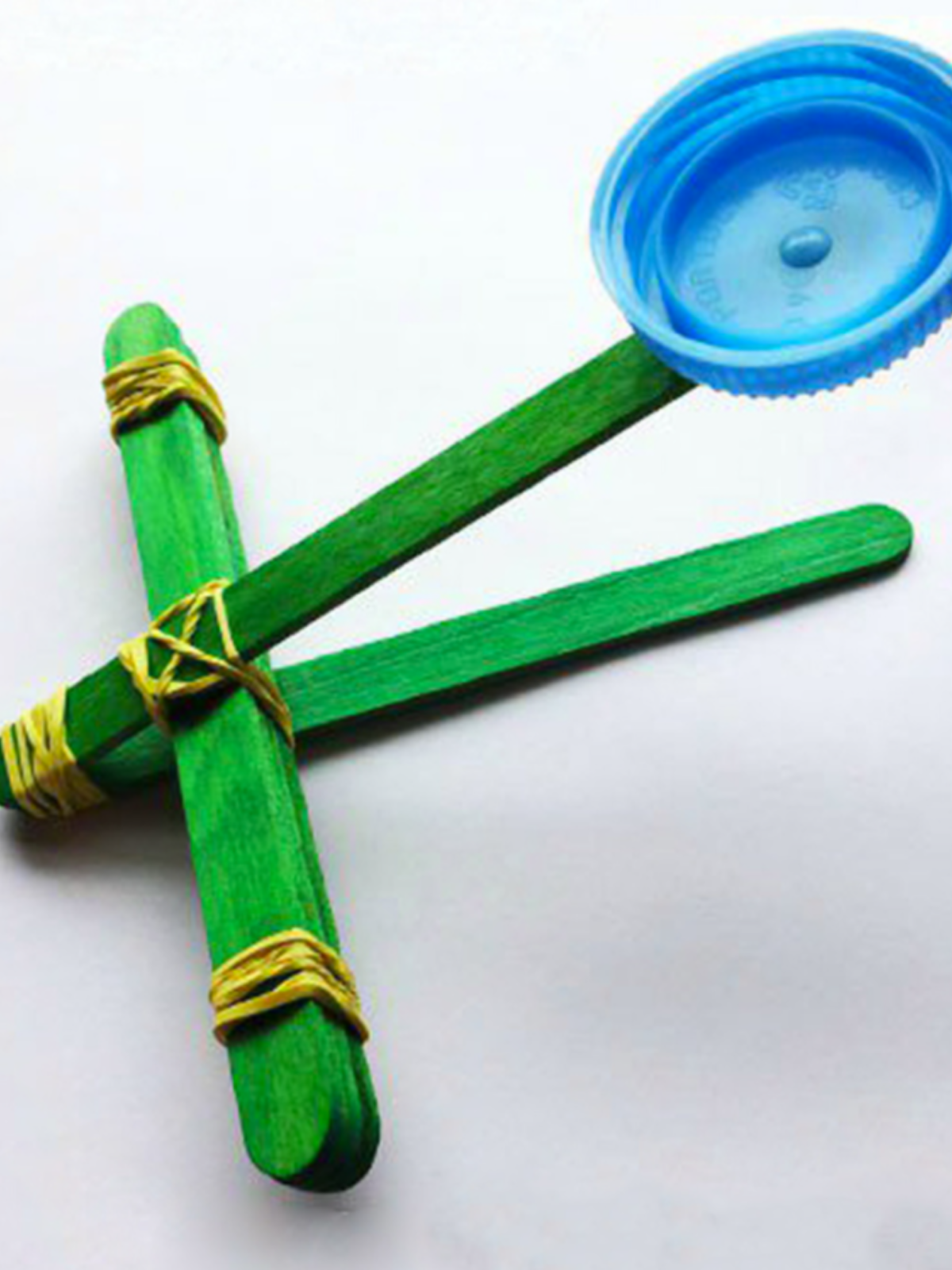 Catapult Craft- green popsicle sticks, rubber bands, and lid to make a catapult on a white background- kids activities blog