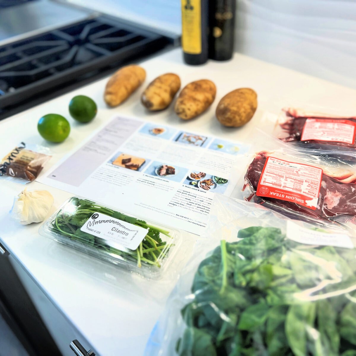 ingredients unpacked from Marley Spoon meal box for steak dinner shown on counter with recipe card - potatoes, limes, spices, garlic, cilantro, spinach and steak with olive oil in the background