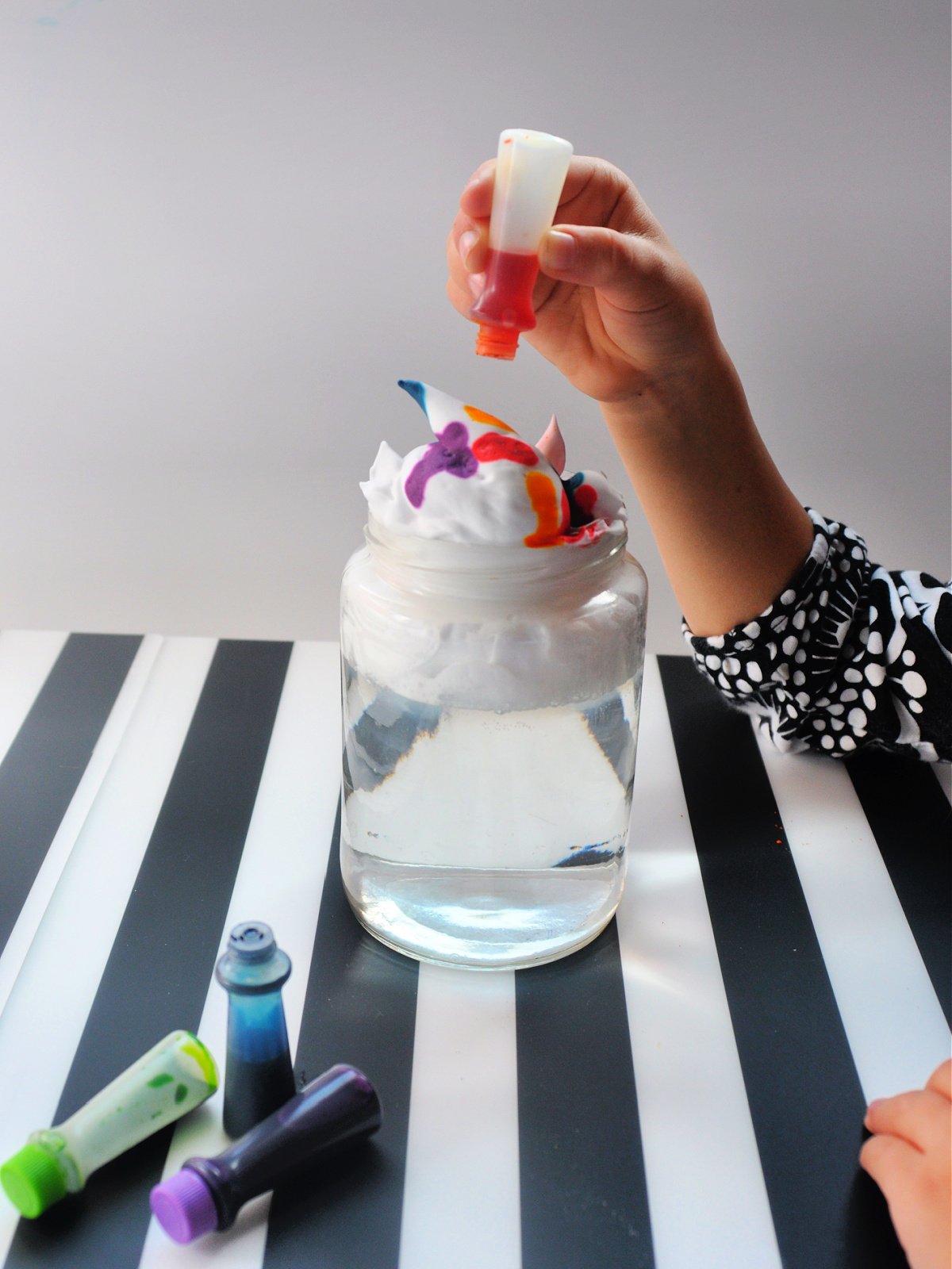 Weather experiment- step 2- hand pouring blue, green, red, and pink food coloring into shaving cream and water- kids activities blog