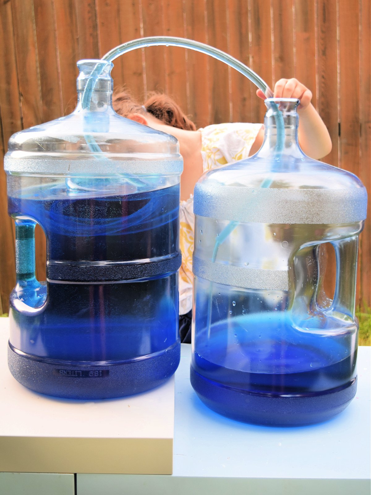 Science Experiment- little girl outside holding clear tubing that is holding and moving water from one container to the next- kids activities blog
