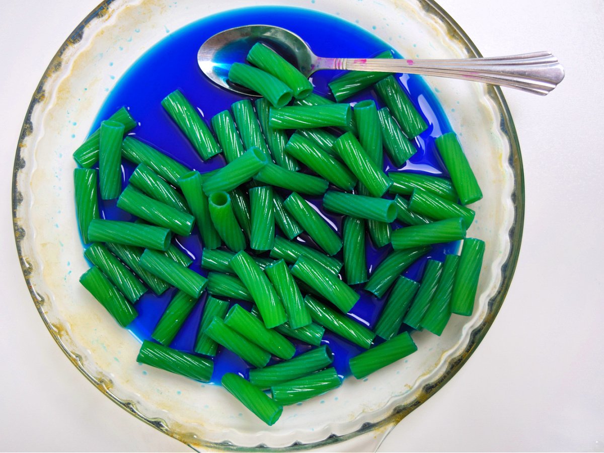 Pasta Fountain Science Experiment- step 1- noodles coated in blue food die in a dish with a spoon - Kids Activities Blog